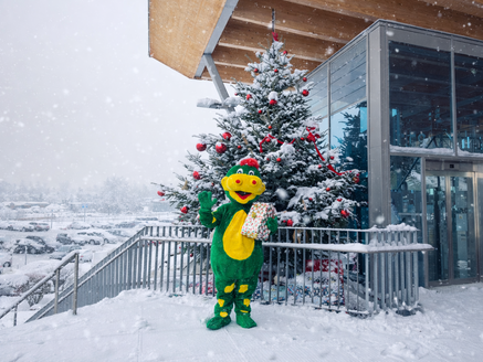 Schmidolin the dragon stands in front of a decorated Christmas tree in the snow holding a gift. Infor: parts of the picture are AI generated | © Schmittenhöhebahn AG