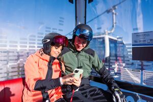 Two skiers in a gondola looking at a phone together. | © mr-offenblende.at