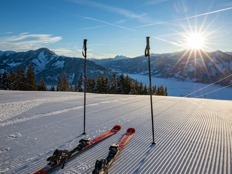 Zwei Skier und Stöcke stehen auf der Piste, Sonnenaufgang über verschneiten Bergen. | © Schmittenhöhebahn AG/Nikolaus Faistauer Photography