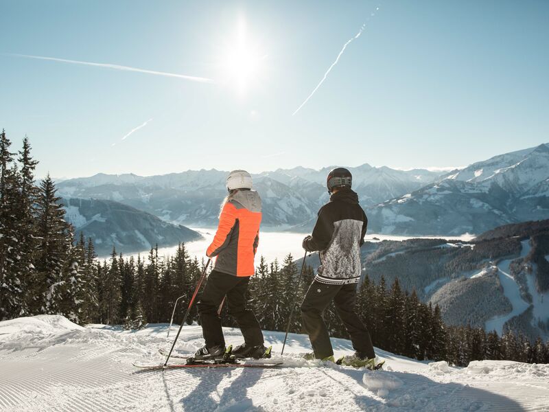 Skifahrer genießen die Aussicht auf verschneite Berge | © Schmittenhöhebahn AG/Punkt und Komma Zwei Skifahrer stehen auf einem Berg und blicken auf die schneebedeckte Landschaft. | © Schmittenhöhebahn AG/Punkt und Komma