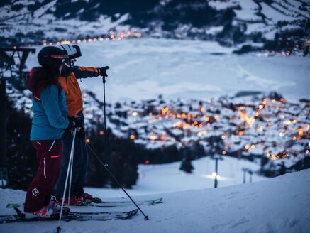 Two skiers on a slope, with a lit village and lake in the background. | © Schmittenhöhebahn AG/Zooom-Productions