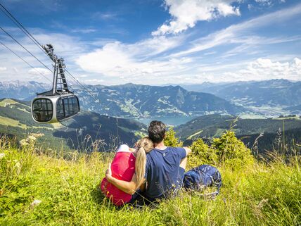 Paar sitzt auf der Schmittenhöhe und blickt auf die umliegende Landschaft mit Seilbahn im Hintergrund. | © Schmittenhöhebahn AG/Max Steinbauer