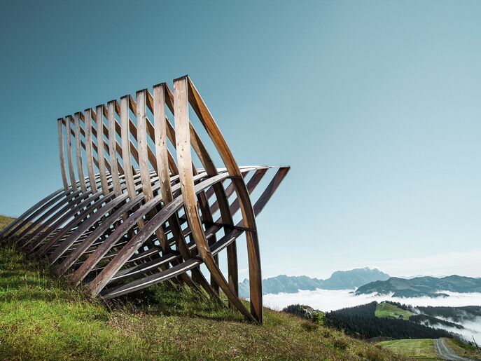 Eine moderne Holzskulptur auf einer Wiese mit Bergen im Hintergrund. | © Schmittenhöhebahn AG/Johannes Felsch 