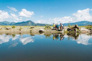 Vier Personen stehen am Ufer eines ruhigen Sees, umgeben von Bergen und blauen Himmel. | © Schmittenhöhebahn AG/mr-offenblende.at