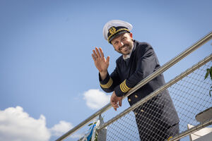 A captain in uniform waves from a railing on a ship. | © Nikolaus Faistauer Photography