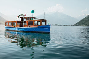 The paddle boat libelle floats on calm waters with mountains in the background. | © Schmittenhöhebahn AG/Johannes Felsch 