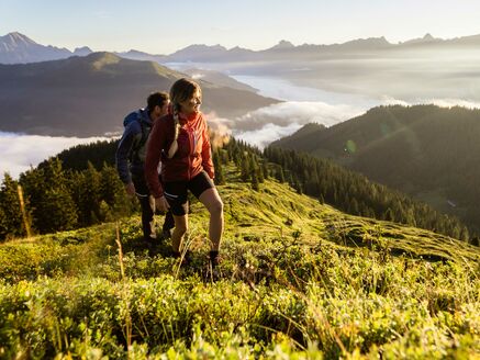 Zwei Personen wandern auf einem Hügel in den Bergen, umgeben von Nebel und einem atemberaubenden Ausblick. | © Schmittenhöhebahn AG/mr-offenblende.at