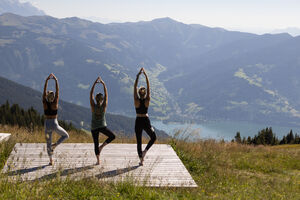 Three people practising yoga on a platform with a mountain view over Lake Zell. | © kuscheiart