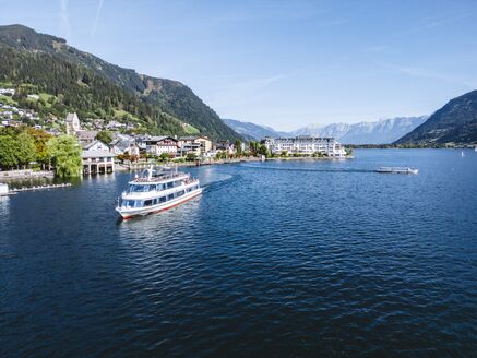 Ein Passagierschiff fährt auf dem Zeller See, umgeben von Grasbergen und einer Stadt am Ufer. | © Alpine Vision