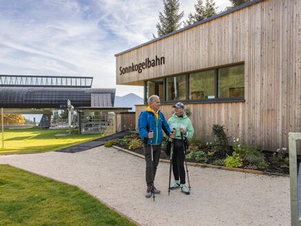 Two hikers with poles in front of the Sonnkogelbahn in a mountainous landscape. | © FotoAtelier Faistauer