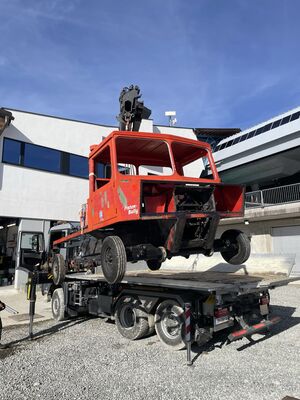 Ausgeschlachtete, rote Pistenraupe mit Aufschrift "Pisten Bully" wird mit einem Kran auf einen Lkw geladen.