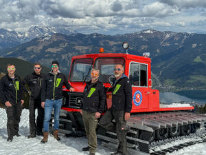 Fünf Männer posieren vor der restaurierten roten Pistenraupe auf einer verschneiden Piste der Schmittenhöhe mit Bergpanorama im Hintergrund.