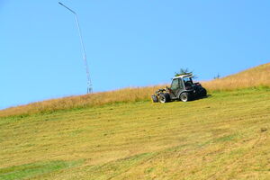 A vehicle mowing grass on a slanted field under a clear sky. | © Schmittenhöhebahn AG